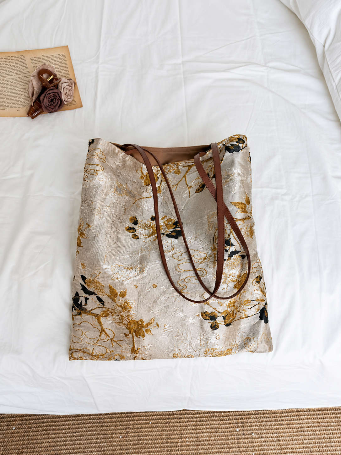 Golden Reverie cream and gold floral jacquard tote with brown handles draped across the front on white bedding.