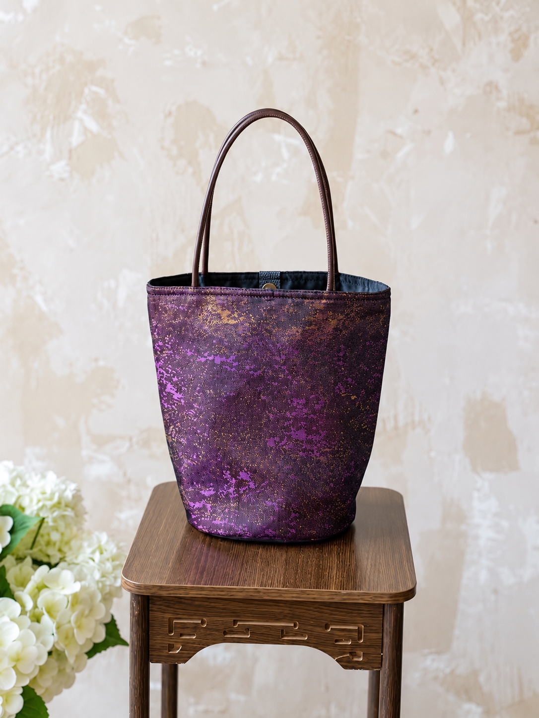 Violet Dusk purple and gold jacquard bucket bag with brown handles, standing on a wooden stool.