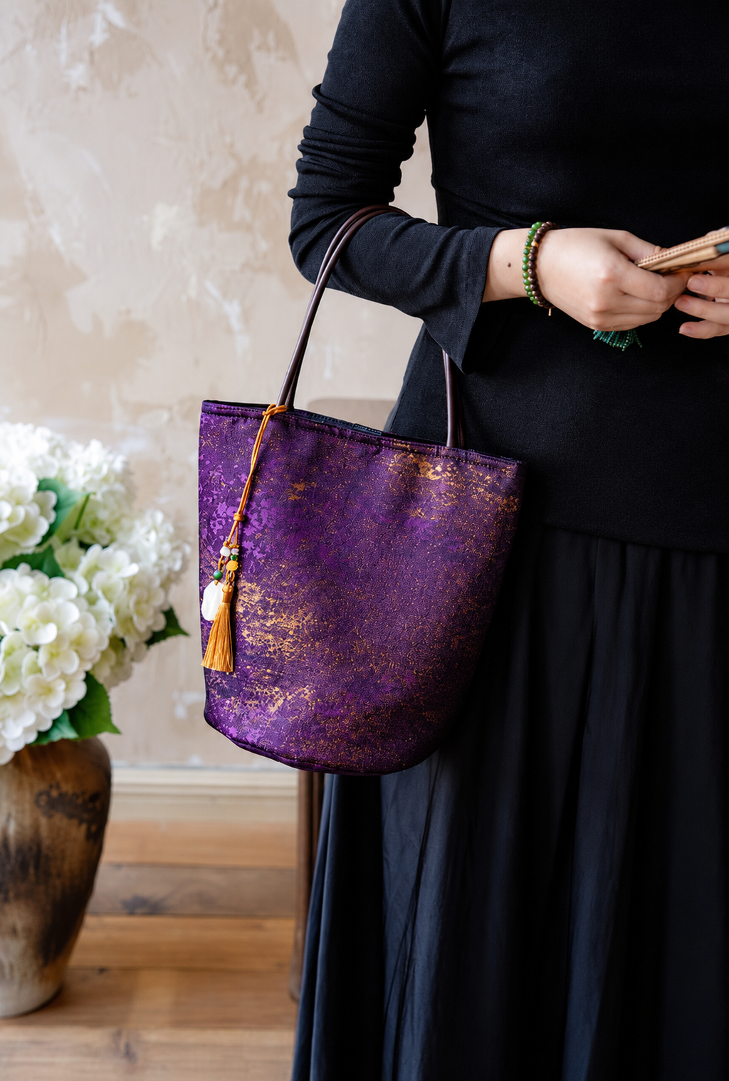 Violet Dusk purple and gold bucket bag carried on the forearm with a black outfit and gold tassel charm.