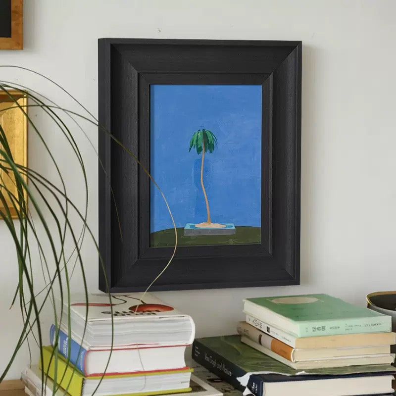 Minimalist framed wall art featuring a stylized green palm tree against a vibrant blue background. The artwork is housed in a
