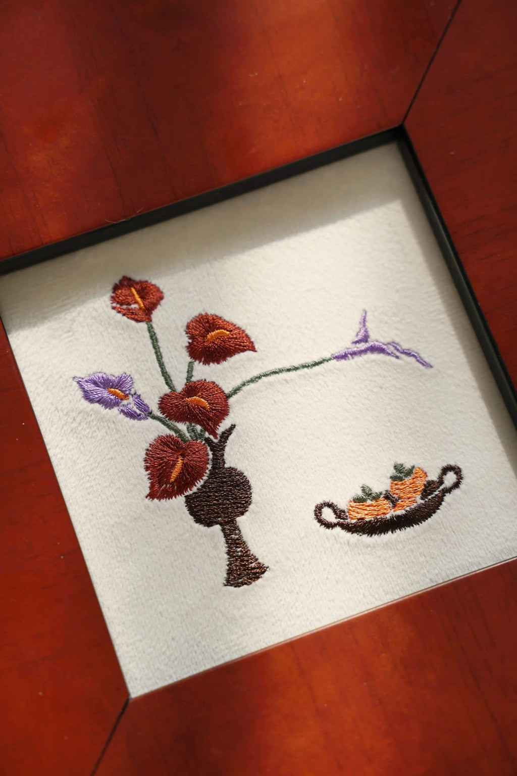 A close-up view of a handcrafted embroidery piece on ivory velvet, featuring a minimalist vase with purple and reddish-brown