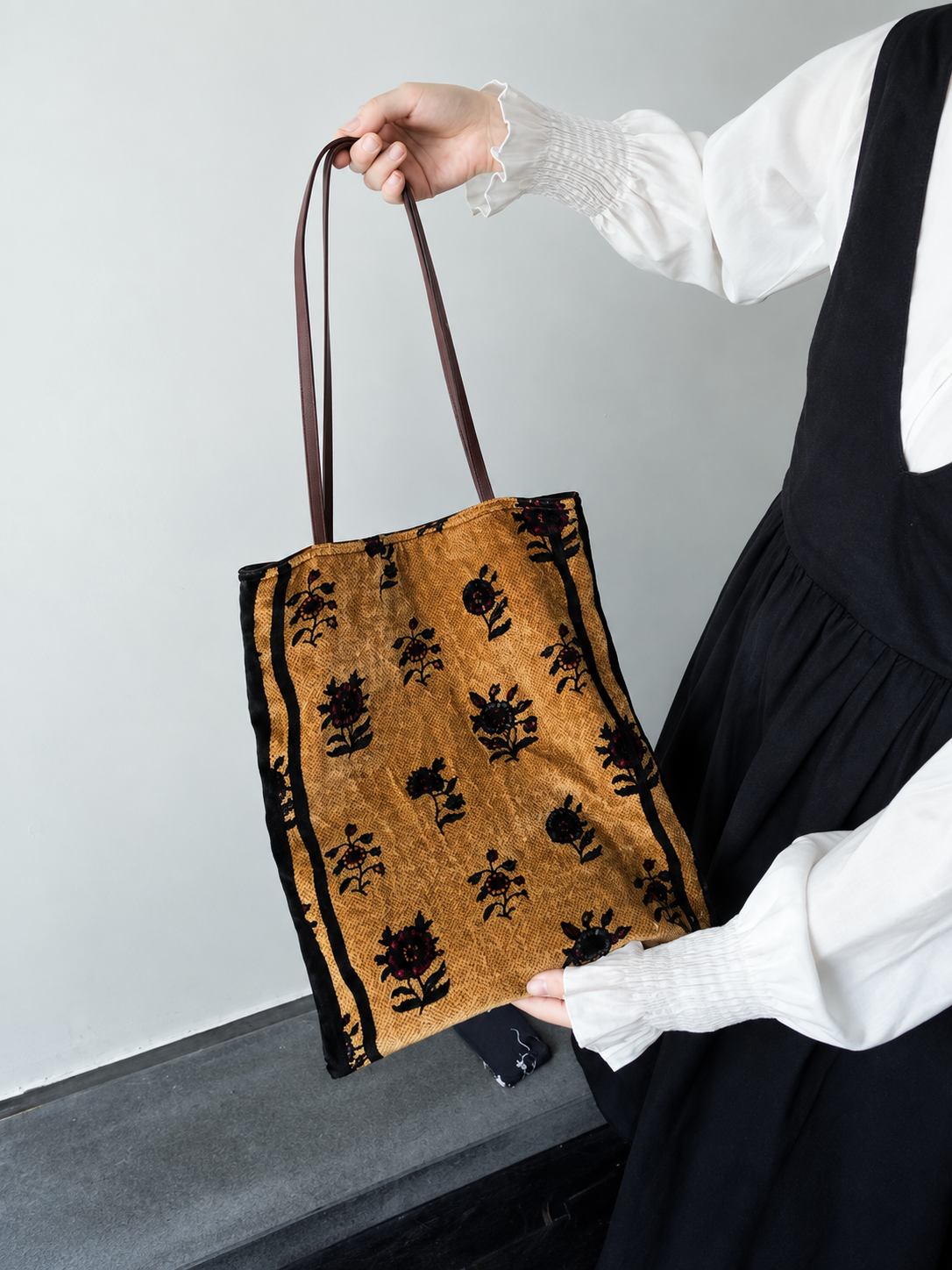 Handheld view of a black and gold floral silk velvet tote bag with long brown shoulder straps.