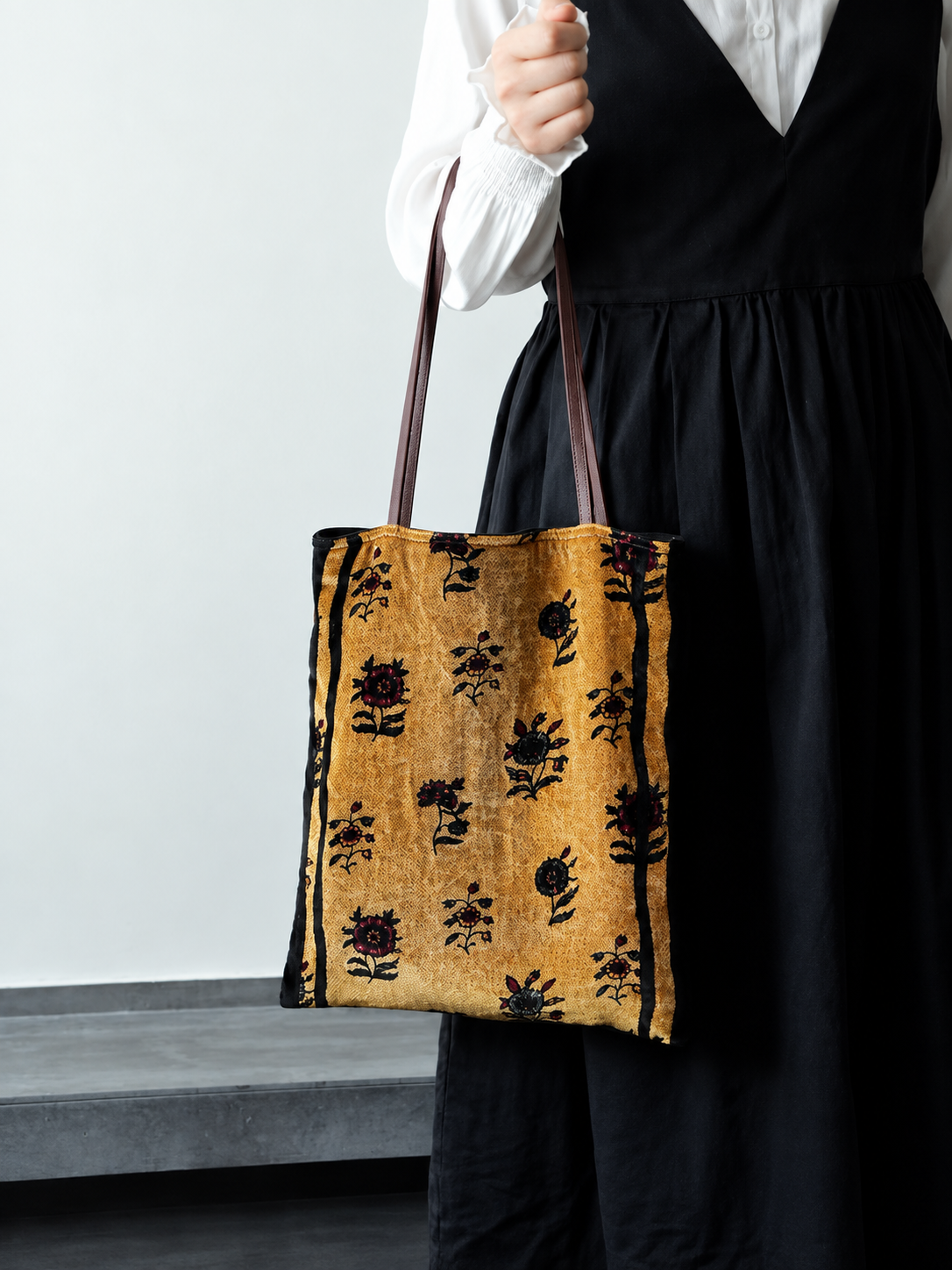 Golden ochre silk velvet floral tote bag carried beside a black dress, showing its rectangular shape.