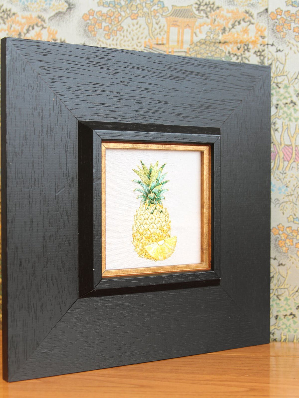 Angled tabletop view of framed golden pineapple embroidery