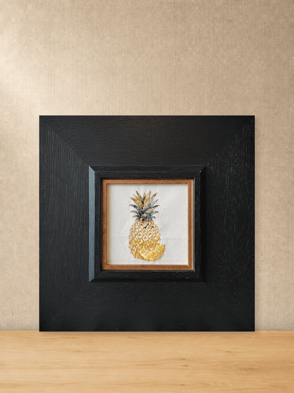 Golden pineapple embroidery in a square black wood frame, front view