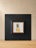 Golden pineapple embroidery in a square black wood frame, front view