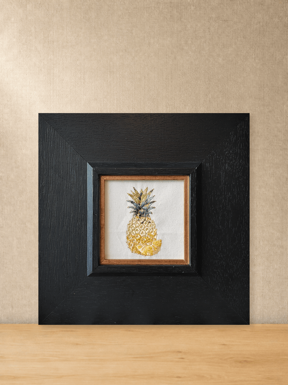 Golden pineapple embroidery in a square black wood frame, front view