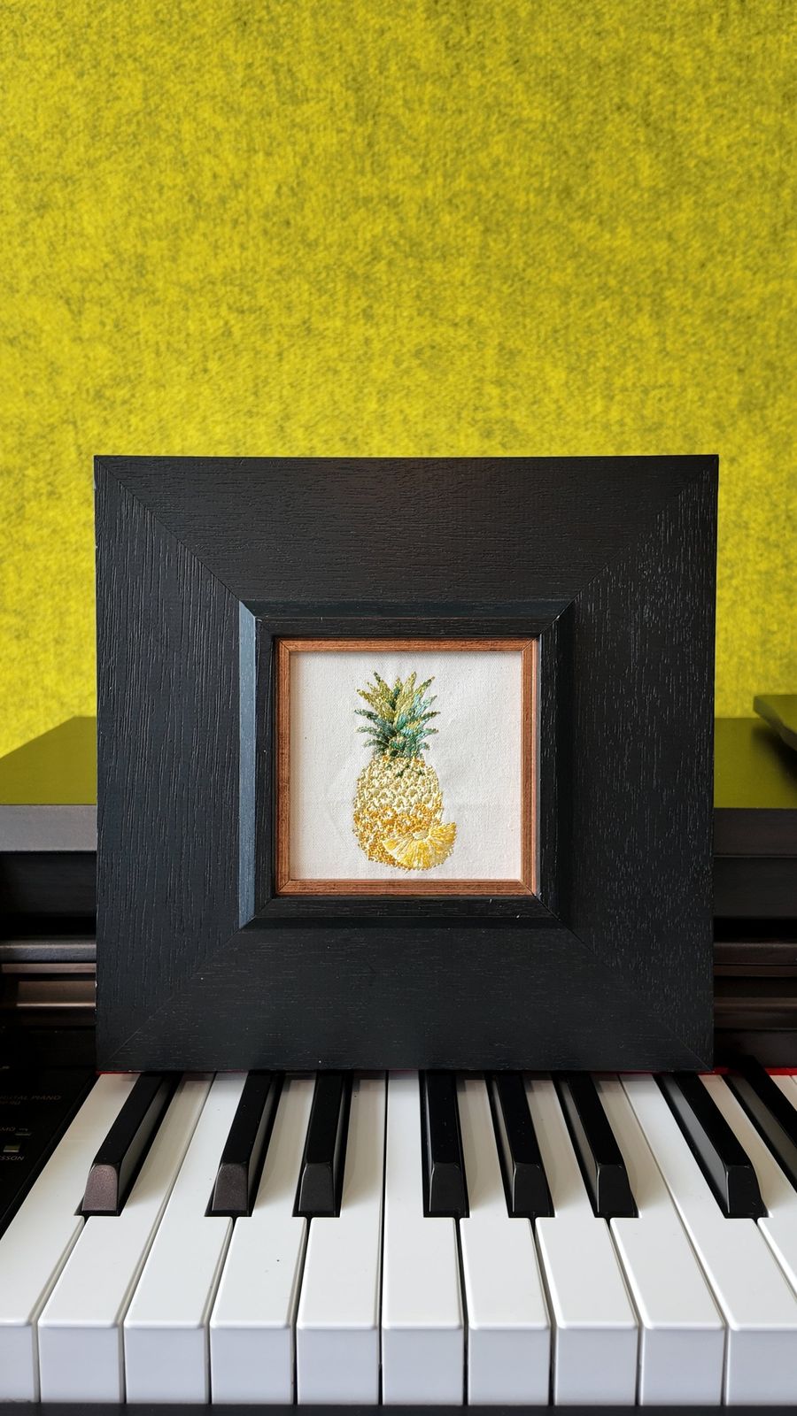 Square pineapple embroidery wall art displayed on a piano