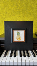 Square pineapple embroidery wall art displayed on a piano
