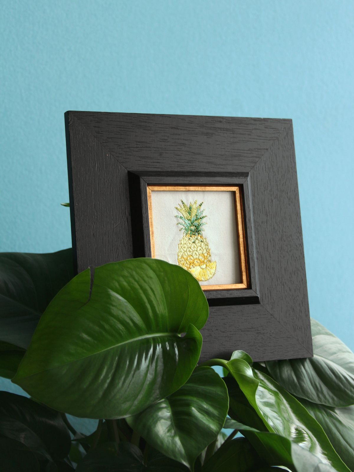 Framed golden pineapple embroidery displayed among green houseplants