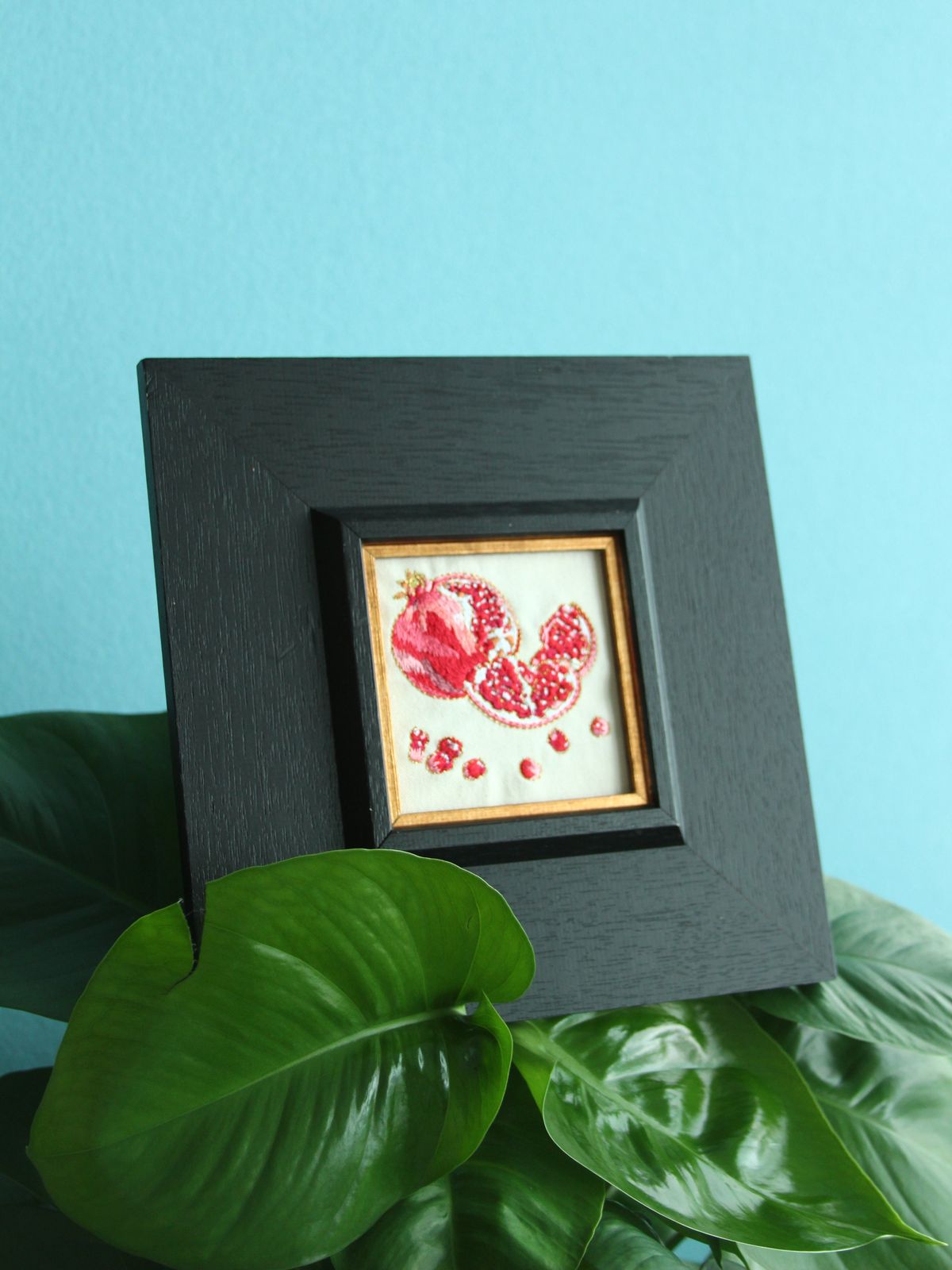 Framed pomegranate embroidery displayed among green houseplants