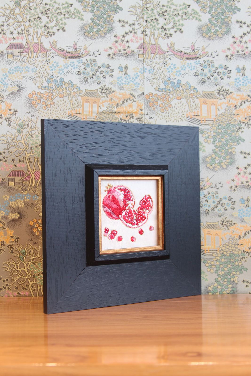 Black framed pomegranate embroidery displayed on a tabletop
