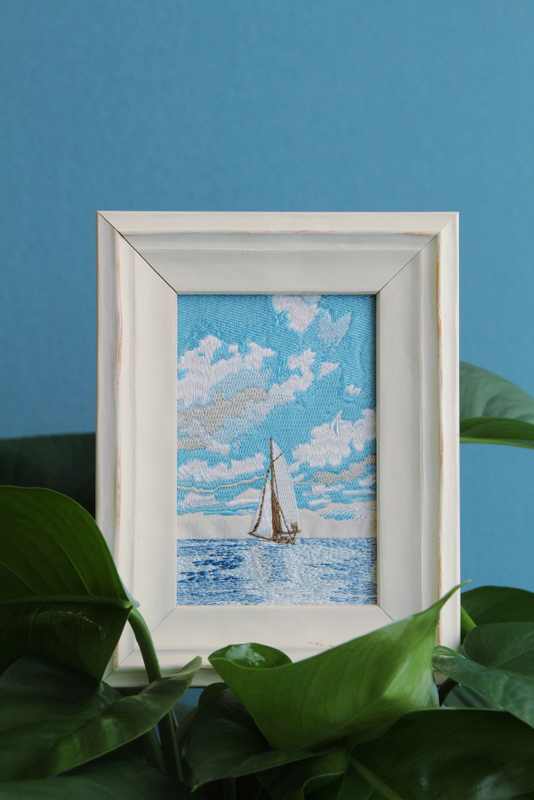 Framed blue sailboat embroidery styled among green houseplants