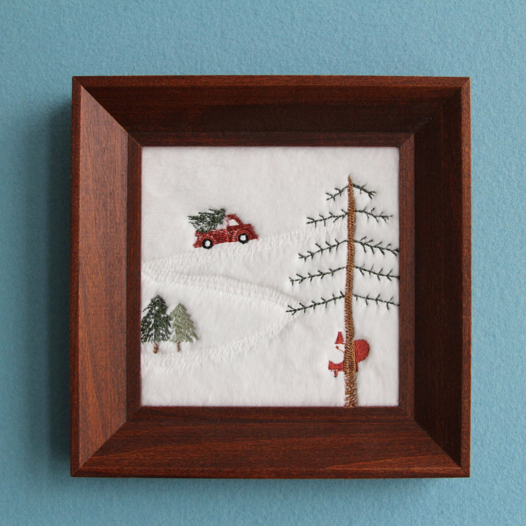Snowy Christmas embroidered wall art in a square brown wood frame, front view