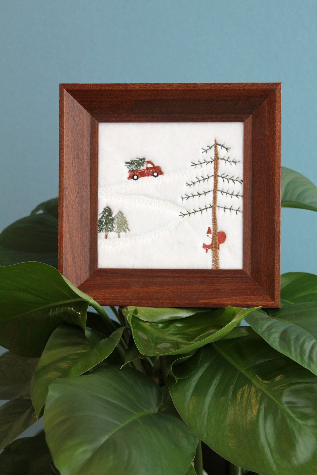 Framed Snowy Christmas embroidery displayed among green houseplants