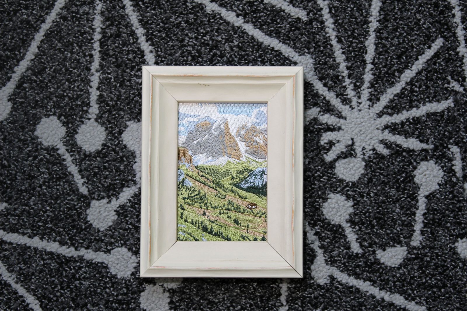 Swiss alpine landscape embroidery in a white wood frame shown in a flat lay