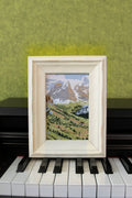 Swiss Alps landscape embroidery in a white wood frame displayed on a piano