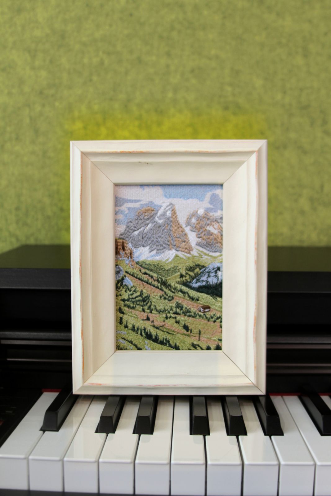 Swiss Alps landscape embroidery in a white wood frame displayed on a piano
