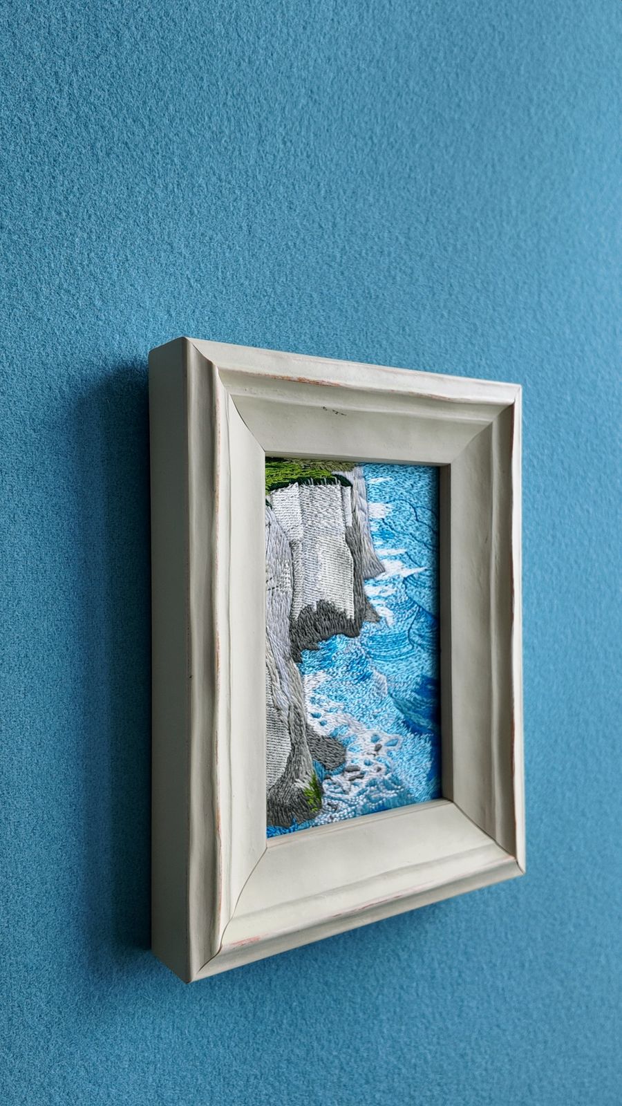 Angled wall view showing the white wooden frame and embroidered seascape